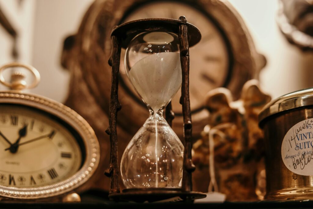 Close-up of a vintage hourglass surrounded by antique clocks in warm lighting.