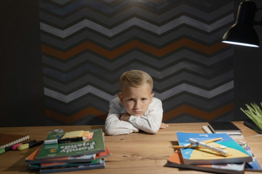 A young boy leaning on a table surrounded by books and coloring materials, looking unhappy with studying.