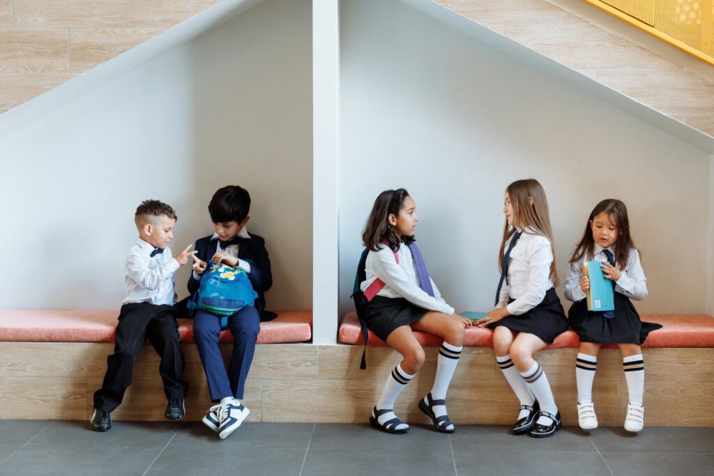 Group of boys and girls in school uniforms talking and sitting indoors on a bench.