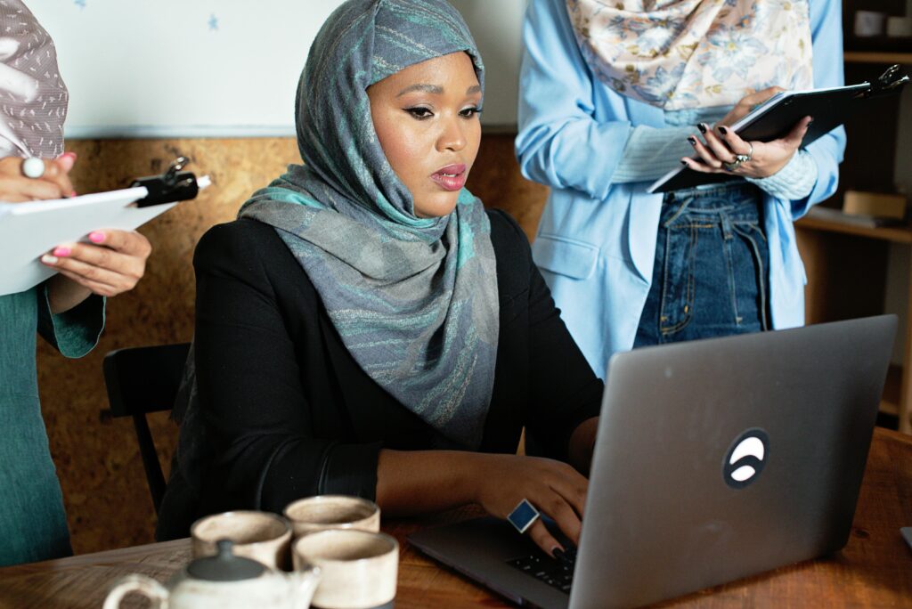 A focused businesswoman wearing a hijab works on her laptop in a collaborative office environment.